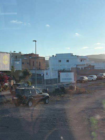 Here's a description of the image:

The image captures a street scene in Puerto del Rosario, Spain, likely taken from inside a vehicle, possibly a bus, due to the presence of a reflection. The view is along a road lined with buildings of varying heights and colors. Several cars are parked along the roadside.

Key elements in the image include:

*   **Buildings:** A mix of residential and commercial buildings.
*   **Signage:** A prominent billboard for "CORORASA" is visible, along with a sign for "Concejalía de Obras". Also, a Lidl sign is visible.
*   **Street Furnishings:** A streetlight is seen.
*   **Vehicles:** Cars are parked on the side of the road, with a car brand model named Jeep.
*   **Vegetation:** Palm trees and flowering plants are present, adding to the streetscape.
*   **Sky:** A bright, blue sky with few clouds forms the backdrop.

Overall, the image depicts a typical street scene in Puerto del Rosario, capturing the mix of buildings, infrastructure, and everyday life in the town.