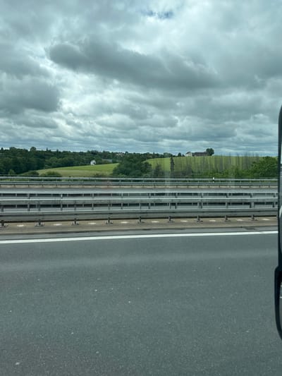 The image shows a view from a vehicle traveling on a highway, likely in the vicinity of Klipphausen, Germany. The foreground is dominated by the asphalt of the road, marked by a white line. Beyond the road, multiple layers of highway barriers are visible, constructed from metal railings and supporting structures. In the background, a lush green landscape unfolds with rolling hills covered in trees and fields. Several buildings, including houses and possibly a church steeple, are scattered across the hills. The sky is overcast with dramatic, grey clouds. The overall scene depicts a typical highway landscape with rural elements in the distance under a cloudy sky. There are no people visible in the image.