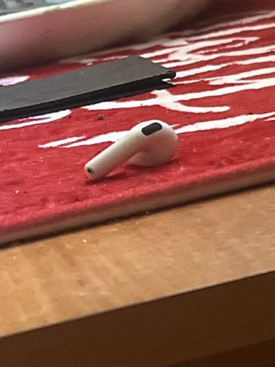 A single white wireless earbud, with a black accent near the tip, rests on a red and white striped surface. A black flat object is positioned to the left of the earbud. The scene is illuminated from above. The red and white surface appears to be a placemat or cloth. The overall setting suggests an indoor environment, possibly a desk or table. There is no indication of the city of Ponebel, Ukraine.