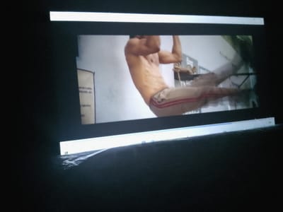 The image is a photograph of a man performing an exercise, likely a pull-up or muscle-up, on a horizontal bar. He is shirtless and appears to be in good physical condition, with visible abdominal muscles. He is wearing athletic shorts with red and white stripes. The background is a light-colored wall, with what appears to be a poster or notice board on the left side. There are also some blurry objects in the background that are difficult to identify. The photograph appears to be taken indoors, possibly in a gym or a similar facility. The image is framed by what look like overhead lights or a projector screen, with one light bar clearly visible at the top and another at the bottom. The lighting suggests a dimly lit environment where the projector or screen is the primary light source. There is no information in the image to suggest a location in Azume, Nigeria.