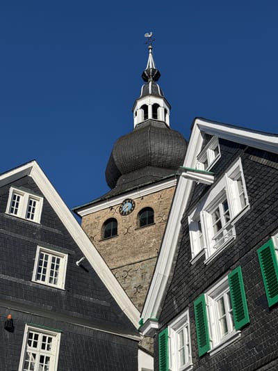 The image depicts a low-angle view of the Klosterkirche, or Monastery Church, in Remscheid, Germany. The spire rises to the upper-left portion of the image. The clock tower features a clock face and arched windows. Dark shingled roofs adjoin the tower on either side of the image with white window frames and green shutters. The sky is clear and bright blue.