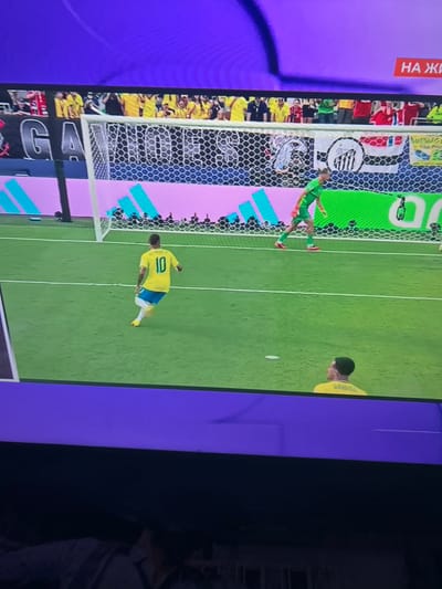This is a photograph of a television screen displaying a soccer match. The screen shows a football field with two players visible. One player, wearing a yellow jersey with the number 10 on the back, is in the foreground, facing away from the camera and towards the goal. Another player, a goalkeeper in a green jersey, is further back on the field, near the goal. Spectators are visible in the background behind a fence. There are also banners and advertising visible around the stadium. The purple border of the television screen is visible at the top and bottom of the image. The location of Ognianovo, Bulgaria, is not depicted in this image; it is a screen capture of a broadcasted event.