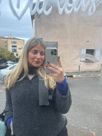 A blonde woman in a grey coat takes a selfie in a mirror. The mirror reflects a street with cars, buildings, and graffiti. The woman holds a silver phone with dark nail polish on her fingers. She wears a ring on her ring finger, and a blue shirt is visible under her coat. The mirror reflects a scene with a muted, overcast sky.