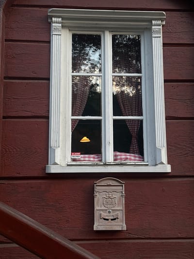The image shows a close-up of a red wooden wall of a building, featuring a white-framed, multi-paned window. Inside the window, red and white checkered curtains are visible, along with a yellow light fixture. A small sticker with the word "ALARM" is affixed to the lower right pane of the window. Below the window, mounted on the wooden wall, is an ornate, light-colored mailbox with a lock. To the left of the mailbox, a portion of a dark reddish-brown wooden railing or beam angles across the lower part of the frame. The overall impression is of the exterior of a rustic building. This scene is located in the city of Krásná Lípa, Czechia.