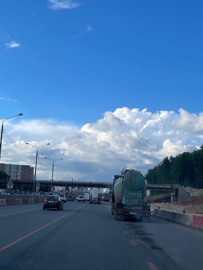 The image depicts a section of road, possibly in Novodrozhzhino, Russia, with several vehicles in a line of traffic. A large, green tanker truck stands out amongst the other cars. The road is marked with a median line and flanked by construction barriers on one side. In the background, a cityscape with apartment buildings is visible, as well as lampposts and overpass structures. The sky is partially covered with a large mass of cumulonimbus clouds. The left side of the image features a clear blue sky.