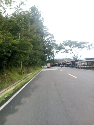 The image depicts a paved, dark gray asphalt road in Deothang, Bhutan, under a bright, overcast sky. The viewpoint is low, looking forward along the road. On the left side, a concrete drainage ditch separates the road from a grassy verge and a steep, densely vegetated hillside covered with green trees, bushes, and some visible reddish blossoms. A utility pole is situated on this slope. The road surface appears damp in patches and is marked with a solid white line on its left edge and two broken yellow lines in its center, indicating two-way traffic. In the mid-ground, a yellow and pink van is visible on the right side of the road, facing away from the viewer. Further down the road, on the right, a cluster of small, rustic buildings with corrugated roofs suggests a market or commercial area. Faint figures, possibly on a motorcycle, are discernible near these structures. A horizontal barrier is partially visible on the far right. No distinct human actions or interactions are clearly observable.