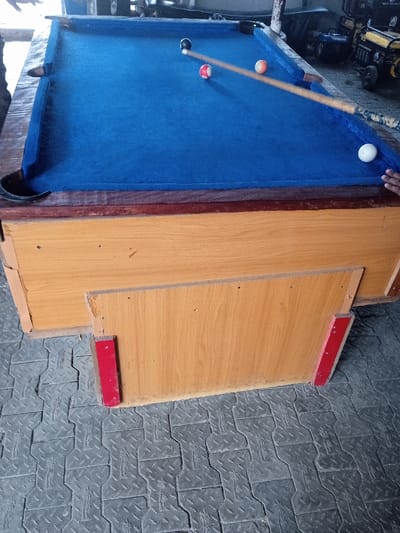 A billiards table with a blue felt top is shown with balls and a cue stick on it. The table's base is wooden and has a partially detached panel with red supports. The table is set on a gray floor with a textured, repeating pattern. The billiards table is in a room with other miscellaneous items.