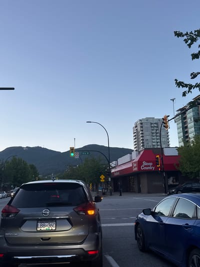 The image depicts a street scene in North Vancouver, Canada, likely during dusk or early evening, with a clear sky and faint light illuminating the city. In the foreground, a dark gray Nissan Rogue SUV is stopped at an intersection, its taillights illuminated. To the right of the SUV, a dark blue sedan is also stopped, its driver's side visible. Across the intersection, a red awning with "Sleep Country" branding extends over the storefront of a commercial building. Behind "Sleep Country," a tall, modern residential building with multiple balconies rises into the sky. Further in the distance, a majestic mountain range forms the backdrop, its slopes covered in dense green trees. A traffic light above the intersection shows a green light, indicating that traffic is permitted to proceed. Another traffic light on the right side of the image is red. A street sign reads "15th ST." The overall atmosphere is calm and urban, with a hint of natural beauty from the mountains.