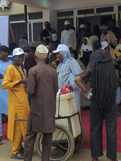 The image depicts a gathering of people in Ungwan Liman, Nigeria. Several individuals are dressed in traditional attire, including a man in a yellow robe and others wearing white caps. One person is eating with a spoon, while another leans over a cart-like structure with wheels. The background shows a building with windows and a red rug on the ground. Some people are observing from the building. Braided hair is visible on one individual.