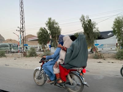 The image depicts a scene in Panjwai, Afghanistan, taken at the intersection of a paved road and a dirt road.  Three figures are riding on a motorcycle. The figure on the right is completely covered in a dark green burqa.  The figure in the middle is also covered in a burqa, but it is a lighter brown color. The figure on the left is a man wearing blue pants and a blue and white patterned shirt. He is driving the motorcycle. The motorcycle is black with red trim, and has a large black seat that can accommodate the three people. There is a large white bus with the word "VIP" in the upper right corner. The bus is parked on the side of the road. There are trees and bushes on the side of the road, including a gas station and a power line pole.  The background features a hill and a clear sky. The scene is captured during the day, with clear lighting and the sun is visible in the sky.
