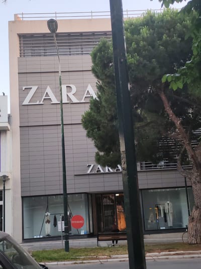 The image shows a Zara store in Alexandroupolis, Greece. The store has a modern facade with the Zara logo prominently displayed. The storefront features large windows displaying mannequins in clothing. A street lamp is visible in front of the store. Trees are also present, with one large tree to the right of the Zara sign. In the foreground, a portion of a car is visible, suggesting the photo was taken from a street. The overall scene depicts a typical urban commercial area.