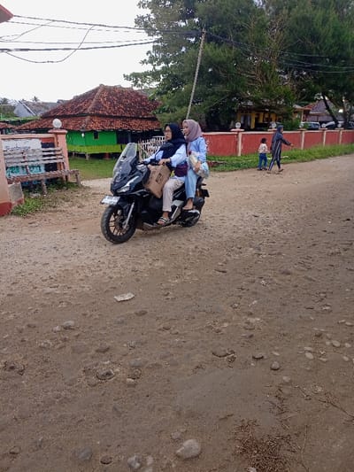 Two women wearing head coverings are riding a black motorcycle down a dirt road in Pameungpeuk, Indonesia (latitude: -7.668, longitude: 107.694).  The woman driving the motorcycle is wearing a black head covering and a long-sleeved black shirt. The passenger is wearing a light blue head covering and a light blue shirt. They both have backpacks on. The motorcycle has a brown box on the back. The road is uneven, with visible tire tracks. There are buildings on the left side of the road, including a building with a red tile roof and a green roof. There is a small child and another woman in the distance. The sky is cloudy.  The motorcyclist is looking straight ahead.  The passenger is looking back at the other woman.
