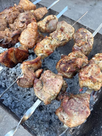 The image shows skewers of grilled meat being cooked over hot coals in an outdoor barbecue pit. The meat appears to be pork or lamb, seasoned with herbs and spices. The coals are gray and white with some red embers visible. The background is a light gray concrete surface. The scene is likely a backyard barbecue in Pokotylivka, Ukraine, given the context provided. There are no people visible in the image.