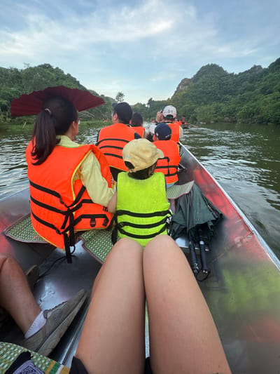 The image depicts a group of people on a boat ride along a river in Yên Sơn Ward, Vietnam. The boat is metallic and carries at least six individuals, all wearing life vests. In the foreground, two individuals' legs are visible, one person wearing shorts and the other whose lower legs are also visible. A person in the front left of the boat wears a yellow shirt and an orange life vest, and holds a dark red umbrella open above their head. Next to them, a child wears a bright yellow life vest and a yellow cap. Other passengers in orange life vests are further back in the boat. The river is calm and surrounded by lush green, tree-covered mountains under a cloudy sky. Two black umbrellas are stowed on the side of the boat. The overall scene suggests a tourist excursion in a scenic natural environment.
