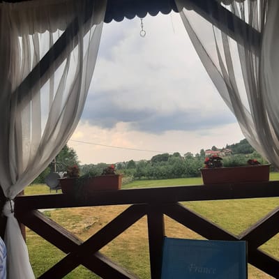 An exterior view from a covered porch, possibly in Dubrivka, Ukraine, overlooks a rural landscape. The foreground features sheer white curtains hanging on either side, framing a view of a wooden railing and then a field. Beyond the field are trees, houses, and a cloudy sky. The wooden railing is a dark brown, X-shaped structure. There are two planter boxes on the railing, one containing red flowers and greenery, and another with more greenery. A blue chair with the word "France" embossed on it sits in the bottom right corner. The sky is overcast with dark clouds.