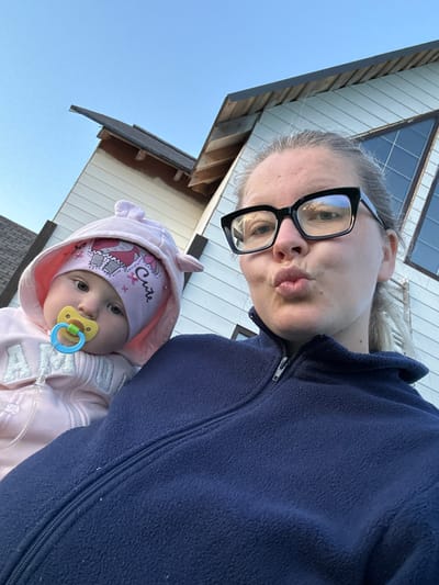 A medium shot captured from a low angle shows a woman and a baby posing for a selfie outdoors. The woman, wearing a dark blue fleece jacket and black-rimmed glasses, puckers her lips. She is holding a baby, who is wearing a pink hooded jacket and a pink beanie with an elephant design and the word "Cute" on it. The baby has a yellow pacifier in its mouth. In the background, a light-colored house with a dark roof and a large triangular window is visible. The sky is clear and blue. The image appears to have been taken in Abushkovo, Russia.