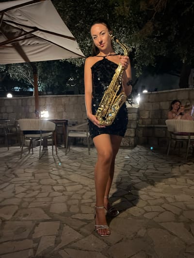 A woman in a black sequined dress and silver strappy heels stands on a stone-paved patio holding a golden saxophone. She is facing forward, looking at the camera with a slight smile. The background shows a restaurant or outdoor seating area with wicker chairs and tables. A large white umbrella is visible to the left, and a stone wall is behind the seating area. Two women are seated at a table in the background, one of whom is looking at her phone. The scene appears to be at night, with artificial lighting. The location is identified as Pečurice, Montenegro.