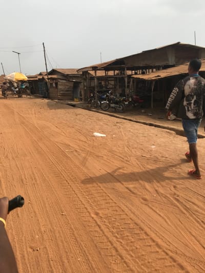 The scene depicts an unpaved, reddish-brown earth road in Ogbese, Nigeria, under an overcast or hazy sky. A row of weathered, single-story structures with corrugated metal roofs and wooden walls lines the right side of the road. Several of these structures appear to be open-fronted stalls, with multiple motorcycles parked in front of them. The road surface exhibits numerous tire tracks and foot impressions. A white plastic bag lies on the ground in the mid-distance.

A male individual is observed from behind, walking away from the camera on the right side of the road. He wears a dark long-sleeved shirt featuring a large white skull graphic on the back, denim shorts, and red flip-flops. His shadow is cast prominently on the ground to his left. In the far left distance, several indistinct figures are gathered near a yellow market umbrella, with additional vehicles parked nearby. A human hand, wearing a yellow bracelet and gripping a black handlebar, is visible in the lower-left foreground, indicating the perspective of a person on a bicycle or motorcycle. Utility poles with overhead wires run along the left side of the road.