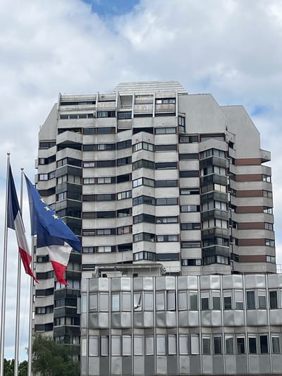 The image shows the exterior of a large, multi-story building in Créteil, France. The building has a modern architectural style with a stepped facade and numerous windows. Two flags, the French flag and the European Union flag, are prominently displayed in the foreground, mounted on flagpoles. The sky is cloudy. The overall scene depicts an urban environment with a focus on the building and the flags.