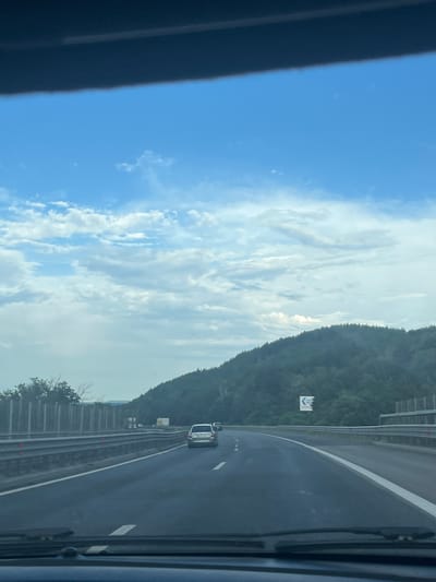 The image depicts a view from the interior of a vehicle traveling on a highway. The road curves to the right, with a green, tree-covered hillside on the right side of the frame. A guardrail runs along the side of the road. In the distance, several cars and a truck are visible on the highway. The sky is blue with scattered white clouds, and the overall lighting suggests it is daytime. There are no visible people or specific landmarks that would definitively place the location in Novi Han, Bulgaria. The scene is a common highway landscape, and without additional context or identifiable features, a precise location cannot be determined from the image alone.