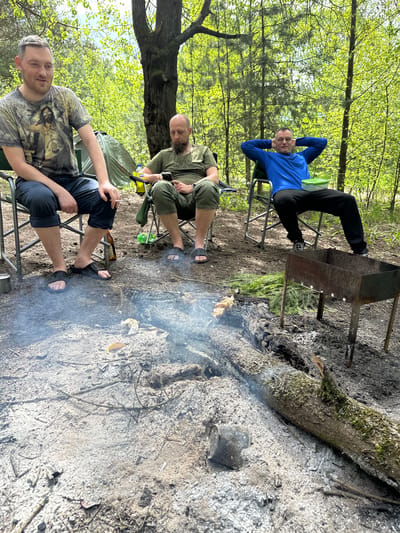 The image shows three men sitting around a makeshift grill in a forest, possibly in the area of Юзуфоўскі сельскі Савет, Belarus. One man, wearing a blue shirt, is reclining in a chair with his hands behind his head. Another man, dressed in an olive-green outfit, is seated in a chair and looking down. The third man, wearing a printed t-shirt, is sitting in the foreground looking toward the camera. A grill with skewers above is visible next to the fire. There is ash covering the ground, indicating a recent fire. A log lies nearby. There is a tent in the background. The lighting is bright, suggesting daytime.