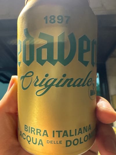 The image shows a close-up, horizontal shot of a person's hand holding a gold-colored aluminum can. The can has green text and graphics on it. The text "1897" is visible on the left side of the can, followed by a large, stylized word that appears to be a brand name, with the letters "P", "E", and "R" clearly discernible. Further along the can, the word "Originale" is written in a flowing script. On the right side of the can, there is text in a more standard font, reading "BIRRA ITALIANA ACQUA DELLE DOLOM". The lighting suggests an indoor setting, possibly with artificial light, and the image is slightly blurry, indicating movement or a shallow depth of field. The person's fingers are gently holding the can, with the skin texture and nails visible. There are no obvious environmental cues to suggest the specific time of day or weather. The overall impression is a casual, candid photograph of a product.