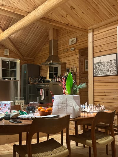 A rustic, wood-paneled kitchen is visible, with a high, angled ceiling supported by exposed beams. Two wooden chairs with woven seats are on the right side of the frame, adjacent to a table cluttered with various items. These include shopping bags, fruit, containers, and other objects. The kitchen appliances, like a stainless steel stove and refrigerator, are centrally located. Several decorative items adorn the walls, including framed pictures and a decorative plate.