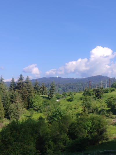 This image presents a serene, sunlit mountainous landscape, characteristic of regions like Momchilovtsi, Bulgaria. The scene is dominated by undulating hills covered in dense greenery under a vast, bright blue sky scattered with fluffy white cumulus clouds.

In the foreground and mid-ground, a rich tapestry of green is visible, featuring numerous coniferous trees, such as firs and spruces, alongside various deciduous bushes and grassy slopes. The vegetation appears vibrant and healthy, indicative of a lush natural environment.

Across the mid-ground, several large power transmission towers (pylons) with multiple high-voltage lines traverse the landscape, introducing an element of human infrastructure into the natural setting. These pylons are visible in varying shades, some appearing reddish-brown, others a lighter metallic gray.

Further in the distance, a prominent forested mountain ridge stretches horizontally. On its peak, two distinct white, dome-shaped structures are discernible, suggestive of radomes or observatory buildings. One large dome is clearly visible on the left side of the ridge, with a smaller one further to the right.

The time of day appears to be midday or early afternoon, given the bright, direct sunlight illuminating the entire scene. The weather is clear and sunny with pleasant cloud cover. No specific activity or event is taking place, and there are no people visible. The image conveys a sense of peaceful natural beauty with subtle indications of human development. There is no visible text in the media file.
