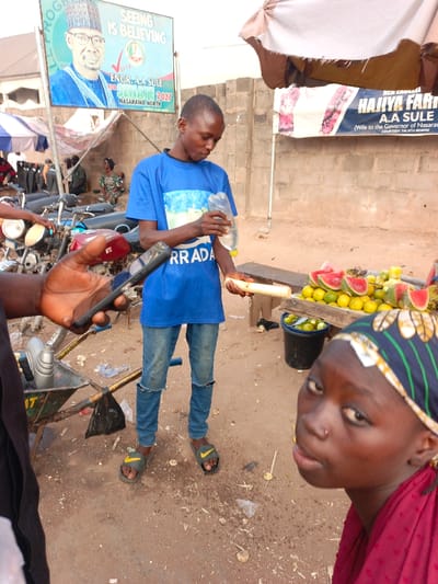 A young man in a blue shirt and jeans stands near a fruit stand. He pours liquid from a bottle onto a peeled sugarcane stalk. Slices of watermelon and oranges are displayed on the stand. A young woman is visible in the foreground. In the background, there are two political posters promoting Engr. A. A. Sule for Senate in 2027 and Hajiya Fatima A. A. Sule, wife to the Governor of Nasarawa. Motorcycles and other people are further in the background.
