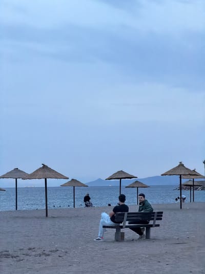 The image shows a beach scene in Glifada, Greece. The sky is overcast. Several straw umbrellas are lined up along the beach near the water. Two men are sitting on a wooden bench. One man is wearing a black shirt and light-colored pants, and the other is wearing a green jacket and dark pants. They appear to be looking at the view. Another person is sitting near the water in the background. The beach is sandy and appears to be sparsely populated.