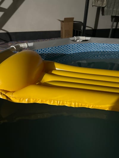 The image shows a close-up of a yellow inflatable mattress partially submerged in dark water. The mattress has a textured surface with raised ridges. To the left of the mattress, a blue and white patterned fabric is visible, possibly part of a pool liner or a covering. In the background, out of focus, are structural elements that appear to be part of an outdoor setting, including metal beams and a cardboard box. The lighting suggests it is either daytime with artificial lighting or nighttime. The scene is still and does not depict any specific activity or event. There is no visible text in the image. The context provided indicates the location as Pîrlița, Moldova.