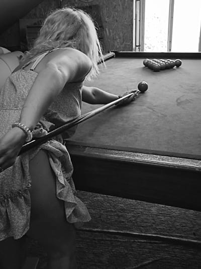 A black and white photograph captures a woman playing billiards in an indoor setting within Терновский округ, Ukraine. The subject, viewed from behind and to the left, has long, light-toned, wavy hair. She wears a light-patterned sleeveless dress with ruffles. Her left arm, adorned with a beaded bracelet, is bent, and her right arm extends forward, gripping a billiard cue. The cue is positioned to strike a cue ball on a dark-felted billiard table. A triangular rack of approximately ten object balls is visible at the opposite end of the table. The room features light-toned, paneled wooden walls and a large, multi-pane window in the background, admitting significant natural light. The floor and lower portion of the wall are constructed of rough-hewn wood. The ceiling appears angled, suggesting an attic or loft space.