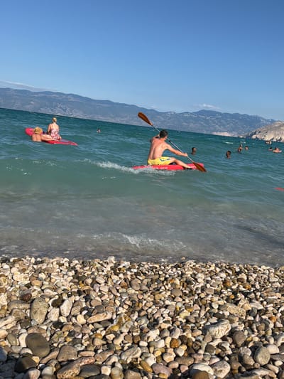 A bright, sunny day at a pebble beach in Općina Baška, Croatia. The water is a clear turquoise, with gentle waves lapping at the shore. In the foreground, smooth, multi-colored pebbles cover the beach. In the middle ground, two people are on a red paddleboard in the water. A man in yellow shorts is paddling a red kayak with an orange paddle. Several other people are visible swimming or floating in the sea further out. In the background, a rugged mountain range with sparse vegetation dominates the horizon under a clear blue sky.