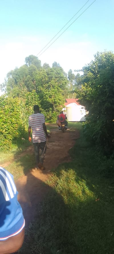 The image shows a dirt path in Isukha ICHINA, Kenya (latitude: 0.352, longitude: 34.779). A man wearing a white and red striped shirt and blue jeans walks on the path, holding a stick. He is walking away from the camera, and his back is to the viewer. In the background, a man on a motorcycle is riding down the path. The motorcycle is facing the camera. There is a white building with a red roof to the right of the path. The building is surrounded by green grass and trees. The sky is blue and clear. The image was taken during the day, with the sun shining brightly.