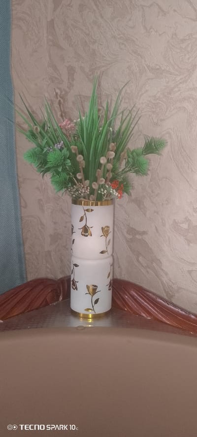 A vertical, medium shot captures a decorative vase filled with artificial greenery and small floral accents. The vase is white with a frosted finish and features a repeating pattern of gold roses. It is positioned on a reflective surface, possibly a table, with a wooden, carved decorative element to its right and a blurred blue curtain to its left. The background is a textured wallpaper with a marbled pattern in shades of beige and tan. The lighting suggests an indoor setting, possibly during the day, as there are no strong shadows indicating artificial light sources. The overall mood is serene and decorative, with no people or discernible activity. The visible text at the bottom of the image reads "TECNO SPARK 10c". The location context is Kliuchi, Russia, though this cannot be determined from the visual information alone.