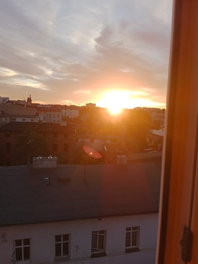 A sunset is depicted over a cityscape in Bydgoszcz, Poland, viewed from a high vantage point. The sun, a bright orange orb, is positioned in the center of the frame, casting long rays of light across the sky and illuminating the buildings below. The sky is a gradient of warm colors, from pale yellow near the sun to soft pink and gray higher up. Wispy clouds are scattered across the sky.

The foreground features the dark, flat roof of a building, with some vents and a small satellite dish visible. Below this roof, a white building with several windows is partially visible. In the middle ground, a cluster of older, multi-story brick buildings with red-tiled roofs are densely packed together. Some of these buildings have visible windows and architectural details. Trees are interspersed among the buildings, adding patches of green to the urban landscape.

In the background, the skyline of Bydgoszcz is visible, with more buildings stretching towards the horizon. A tall, slender chimney or tower rises from one of the buildings in the mid-distance. The overall impression is one of a residential or mixed-use area within the city, observed during the golden hour. The right side of the image is dominated by a blurred, warm-toned vertical element, likely a window frame or wall, which adds a sense of depth and perspective. No people are visible in the frame.