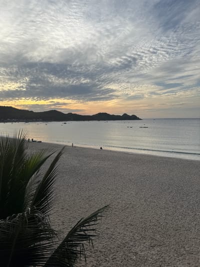 The image depicts a serene beach scene at dusk in Lệ Uyên, Vietnam. The sky is filled with dramatic, textured clouds, ranging from bright white to darker grays, with a warm, golden glow on the horizon where the sun has set. The sea is calm and reflects the muted colors of the sky. In the distance, a chain of dark, silhouetted hills lines the coast. Several small boats are scattered across the water.

The beach itself is composed of fine, light-colored sand. A few individuals are visible on the sand near the water's edge. On the left side of the foreground, the fronds of a dark green palm tree partially obscure the view of the beach and sea, adding a tropical element to the composition. The overall atmosphere is peaceful and tranquil.