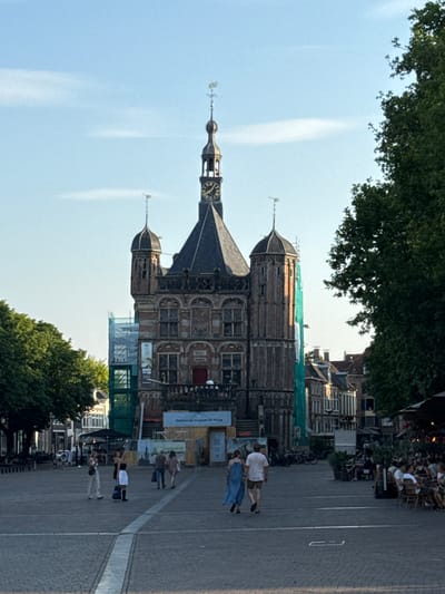 The image depicts a street scene in Deventer, Netherlands, with a prominent building featuring a clock tower. The orientation of the image is sideways. People are visible walking and standing on a cobblestone street and in an outdoor seating area. Some individuals are casually dressed, suggesting a pleasant day. The weather appears to be clear with a blue sky and some wispy clouds. The time of day seems to be late afternoon or early evening, judging by the lighting.
The main building is an architectural landmark, possibly a historic town hall or similar public structure, with intricate brickwork and decorative elements. Scaffolding and green netting are present on parts of the building, indicating ongoing renovation or construction work. Several bicycles are parked along the street. In the background, there are more buildings, some with awnings and outdoor seating for cafes or restaurants, suggesting a lively urban environment.
Some text is visible on a banner or sign in the middle ground, reading "Museum" and possibly part of a longer phrase in Dutch. Another sign on a construction barrier also has some text, though it is not clearly legible. The overall atmosphere appears relaxed and social, with people enjoying the public space.
