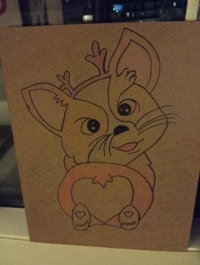 A hand-drawn illustration of a cartoon deer-like creature with large ears and antlers is centered on a piece of brown paper. The creature is colored in shades of orange and pink, with black outlines. It has large, round eyes, a small nose, and a smiling mouth. Its paws are holding a heart shape. The paper is leaning against a window, with a blurred city skyline visible through the glass. The image is lit by artificial light, casting soft shadows. The drawing itself is the primary subject, with the background providing context of its placement. There are no people or discernible interactions present in the scene. The location in Anderlecht, Belgium, is not visually evident within the image itself.