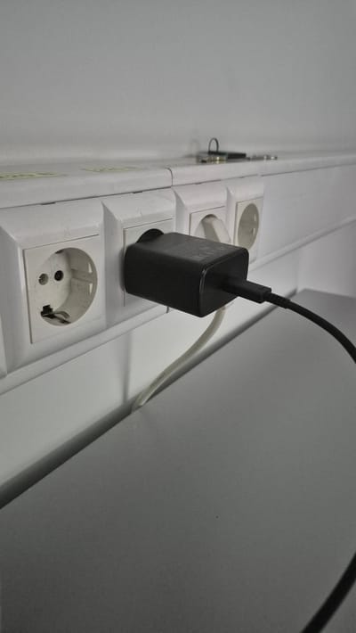 The image shows a close-up view of a white power strip with multiple European-style outlets. A black power adapter, likely for a phone or other electronic device, is plugged into one of the outlets. A black cable extends from the adapter and disappears out of frame. A white cable is also visible, running from another outlet and alongside the power strip. The power strip is mounted on a white wall, which appears to be indoors. The lighting suggests it is daytime, but there are no windows or external cues to indicate the time of day. The overall scene is simple and functional, focusing on the electrical outlets and the charging adapter. The power strip has some visible markings or text on its surface, but it is not clear enough to be legible. The context suggests a domestic or office environment where devices are being charged.