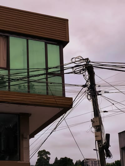 In the city of Charnali, Georgia, an outdoor shot captures a street scene dominated by a utility pole heavily laden with electrical wires and telecommunication cables. The pole, positioned to the right of the frame, is a dark, weathered cylinder. Attached to it are two square, cream-colored junction boxes, one above the other, with visible wiring and coiled cables.
To the left of the pole, a portion of a modern building is visible. The building features a section with horizontal, brown siding on its upper level, beneath which is a band of large, green-tinted windows. A lower level of the building, partially obscured, has a darker window.
The sky above is overcast, with a soft, diffused light, suggesting either dawn or dusk. In the background, beyond the building and utility pole, a dense arrangement of trees can be seen, with the suggestion of other structures, possibly more buildings, in the far distance. The overall impression is one of urban infrastructure with a focus on the intricate network of overhead wires. There are no people or animals visible in the image.