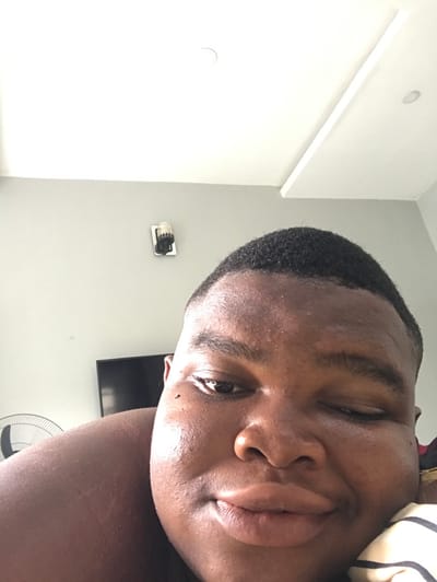 This image depicts a close-up, low-angle selfie of an individual in an indoor residential setting in Langbasa, Nigeria.

**Person:** One individual, appearing to be an adult with a dark complexion, occupies the majority of the frame. The subject's face is prominent, featuring short, dark, closely cropped hair with faded sides. The individual is looking directly towards the camera with a slight smile; their left eye (viewer's right) is partially closed or squinting, while the right eye (viewer's left) is open. The subject's full lips are slightly pursed. A bare shoulder and upper arm are visible on the left side of the frame, indicating the individual is likely shirtless or wearing a garment that exposes the shoulder. The head rests on a pillow with diagonal cream and brown stripes, partially visible in the bottom right corner.

**Environment and Objects:** The background consists of a light grey wall in the mid-section, transitioning to a white ceiling above. On the light grey wall, to the left of the subject's head, a white wall plate with a black and silver wall-mounted device (possibly a sensor or camera) is visible. The white ceiling features a single recessed circular light fixture and a longer, rectangular flush-mounted light panel. In the upper left background, behind the subject's left shoulder, a dark, flat-screen television is partially visible. Further to the left, behind the subject's left arm, a white oscillating fan is partially visible. The lighting appears uniform, suggesting an interior space with ambient or overhead artificial illumination.