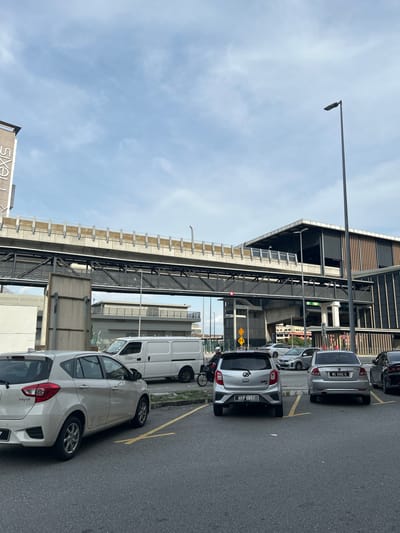 A low-angle, full shot captures a public transportation hub or complex in Subang Jaya City Council, Malaysia. The scene is dominated by an elevated walkway or train line constructed with concrete and metal beams, running horizontally across the mid-ground. Below the elevated structure, a modern building with a facade of vertical wooden slats and dark glass windows is visible. In the foreground, a parking lot with several cars parked is depicted. Two light-colored cars, a hatchback and a sedan, are prominently featured. A white van is parked to the left of the frame. The ground is asphalt, marked with yellow parking lines. A motorcycle with a rider wearing a helmet is moving through the parking lot. The sky above is overcast with scattered clouds, and a streetlamp stands tall on the right side of the image. A building with "nexis" vertically displayed on its side is partially visible on the far left, suggesting the location might be Nexus Bangsar South or a similar commercial complex.