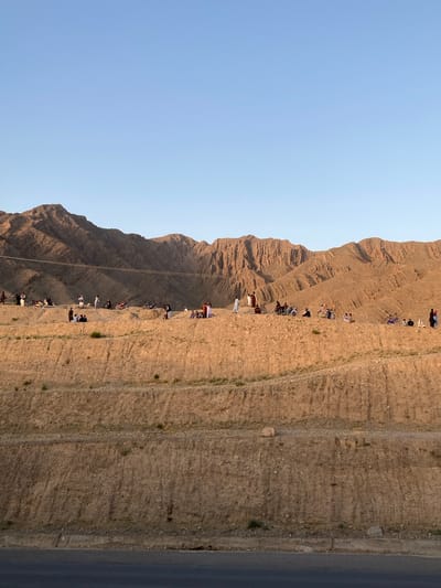 A landscape photo from Kuchlak, Pakistan shows a tan, mountainous region under a light blue sky. A line of people on bicycles and on foot is seen traveling along a narrow path parallel to the mountain face. The path is adjacent to a darker-colored asphalt road. Some people wear white clothing. The ridged texture of the mountain is illuminated by sunlight. Overhead wires are visible along the mountain's slope.
