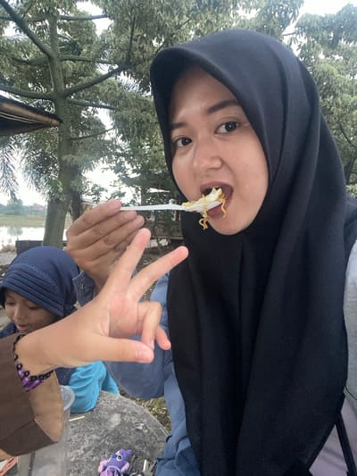 The image displays an outdoor scene in Kalitengah, Indonesia, featuring two individuals and a visible hand. In the right foreground, an adult female wears a black hijab and a light blue top. She is captured mid-action, consuming yellow noodles and a white food item from a white plastic spoon held in her right hand, with her mouth open. Her eyes are directed towards the camera. To the left, partially obscured, a younger individual, consistent with a child, wears a dark blue head covering and a light blue garment; their face is visible, and they appear to be looking downward with a slight smile. A fair-skinned hand, positioned centrally in the foreground and likely belonging to the child, displays a "peace sign" (V-gesture). Both the visible hand and the child's arm wear dark beaded bracelets with purple accents. In the immediate foreground, on a light-colored, rough surface, a small purple My Little Pony figurine and a set of metallic keys are visible. A brown bag and a clear plastic cup are also present. The background consists of dense green foliage from trees, some exhibiting light-colored leaves, under an overcast sky. To the left, a body of water, possibly a pond or flooded rice field, is visible, with distant houses and sparse vegetation on the far bank.