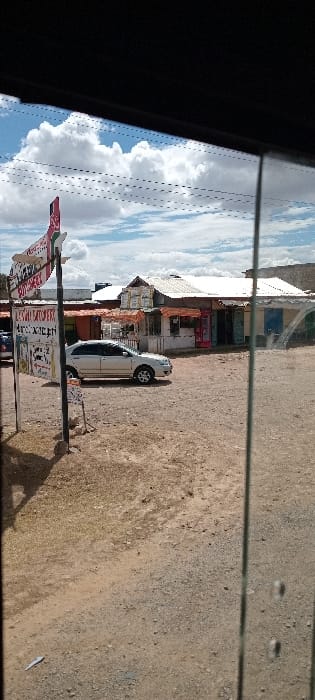 A white car is parked on a dirt lot in front of a row of small buildings with storefronts. A sign with text is positioned in front of the car. The sky is partly cloudy, and the image is viewed through a window, indicated by reflections and the window frame. The scene conveys a sense of rural or small-town life.
