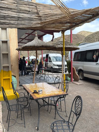 This image shows an outdoor seating area, likely a café or restaurant, located in Zerkten, Morocco. The tables are set with decorative yellow tile tops and metal chairs. Several people are seated and walking around in the background, partially obscured by the thatched-roof umbrellas. A white van is parked nearby. The area is partly shaded by a bamboo structure. The location is possibly near a mountain or rock formation visible in the background.