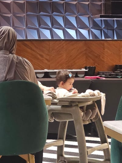 A child with a blue hair clip plays with a stuffed animal seated in a vintage metal high chair. The chair has a light grey frame and a tan-colored tray. A person sits in a green chair nearby, seemingly observing the child. The background is a mix of textures, including a wall with geometric, pyramidal tiles, a wooden surface with grain patterns, and a vertical structure with circular openings.