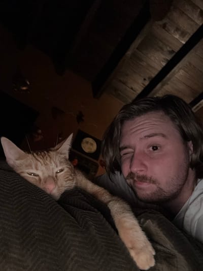 The image depicts a man and an orange cat lying on a dark-colored, possibly corduroy, surface. The man has brown hair and a beard, and is wearing a light-colored shirt. The cat is nestled close to the man. In the background, there is a dark room with a wooden ceiling and a few objects on a table, including a circular item which may be a decorative plate. There is no information available in the image to associate it with the city of Cañada de Madero, Mexico.
