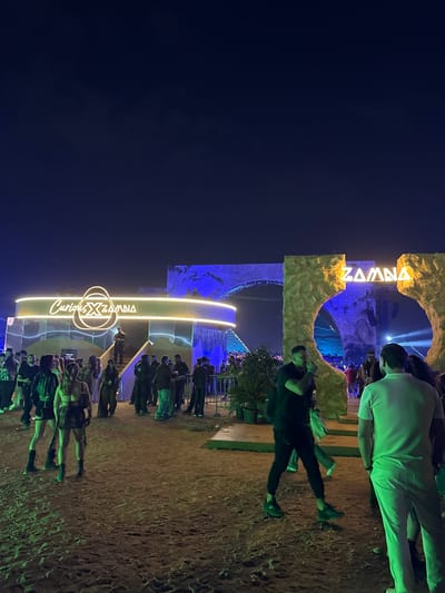 The image depicts a nighttime scene at what appears to be an event or festival entrance, possibly located in Giza, Egypt, based on the word "ZAMNA" visible in the scene's architecture. People are milling around, some walking towards the entrance archway. The entrance itself is a large, ornate structure with illuminated signage, possibly indicating the event's name or theme. The lighting is a mix of artificial glows emanating from the event entrance and overhead ambient lighting. The ground appears to be dirt. Some individuals can be seen directly in front of the entrance while others are slightly further back, forming a loose crowd. The sky is dark, suggesting it is late in the evening or night.